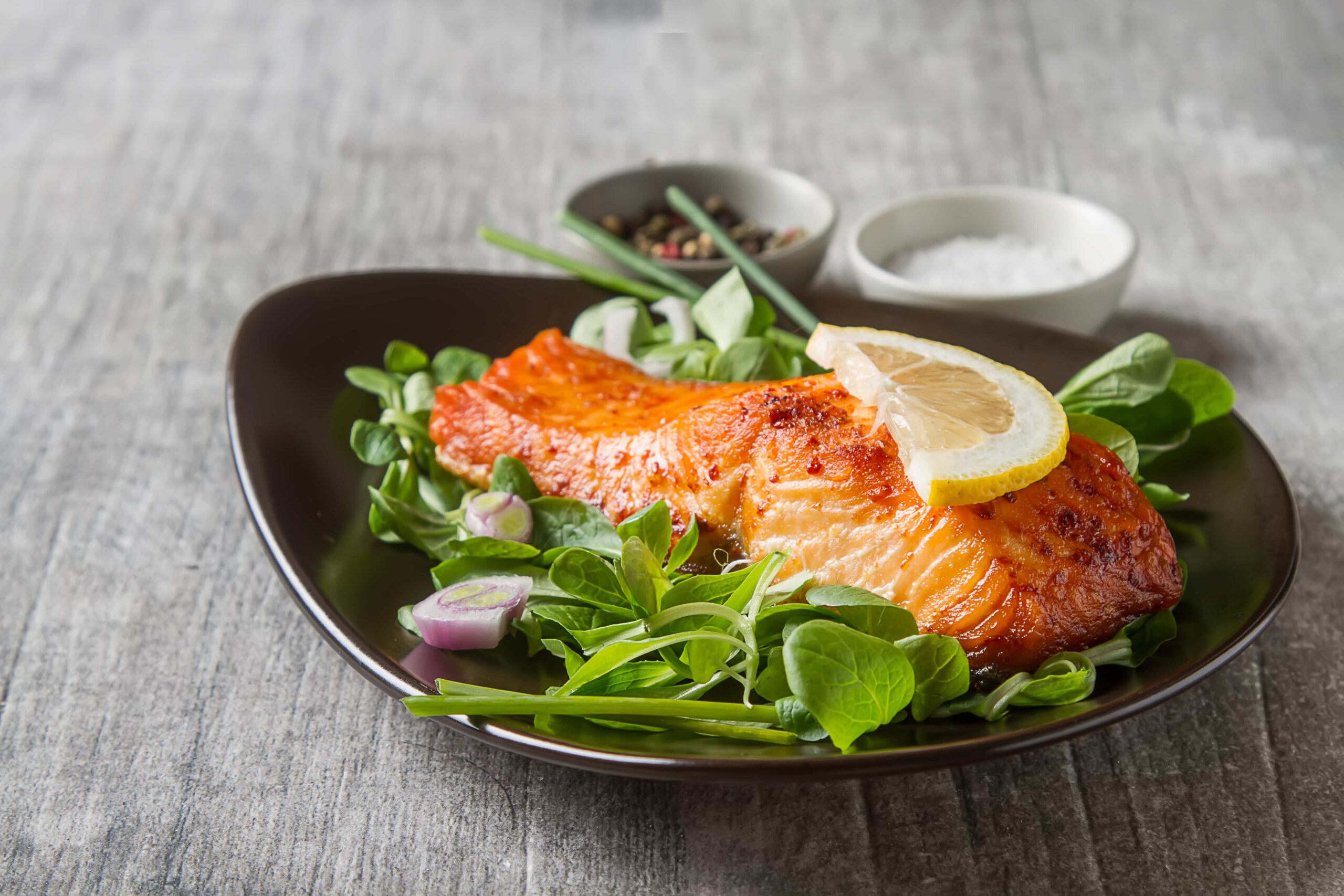 Baked Scottish salmon sits on a black plate on top of a bed of lettuce, topped with a lemon slice. 