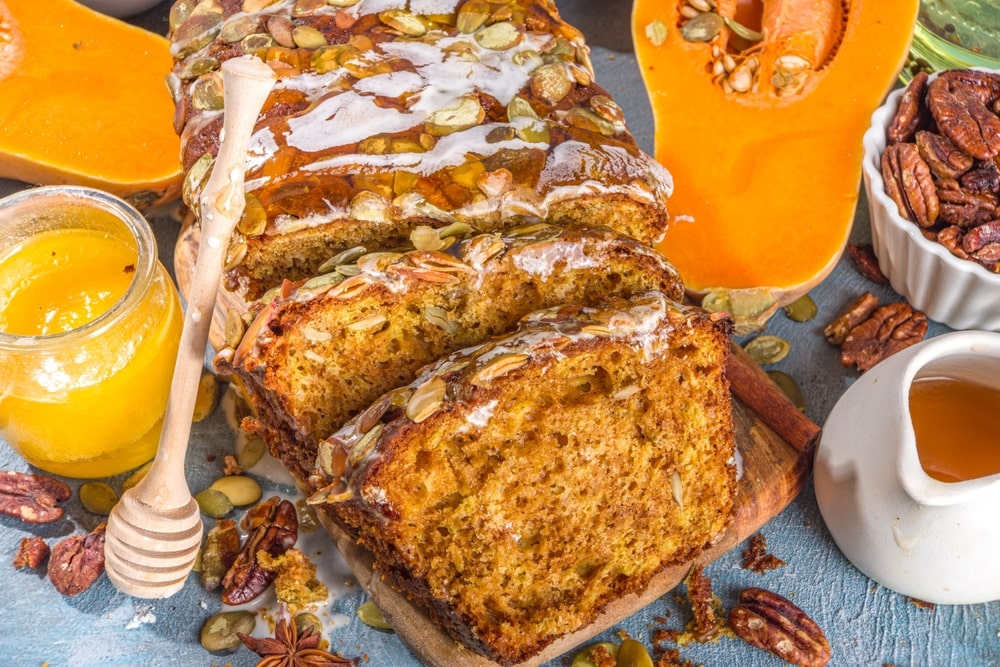 Loaf of gluten-free pumpkin bread surrounded by seeds, pecans, a honey dipper, and cut squash on a table