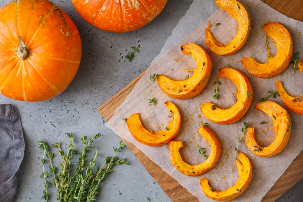 Baked pumpkin slices covered with thyme on a wooden board over a table. Two whole pumpkins are off to the side.