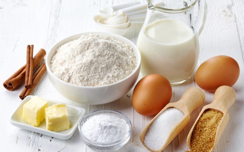 A group of ingredients used for baking, including a bowl of flour, a pitcher of milk, eggs, sugar, and butter.