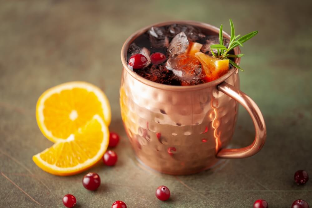 A Moscow mule mocktail in a copper mug with cranberries, orange slices, and a rosemary sprig on top