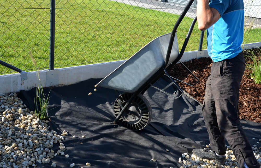 Mulch delivery team member unloads stones from a wheelbarrow onto a tarp in a garden bed
