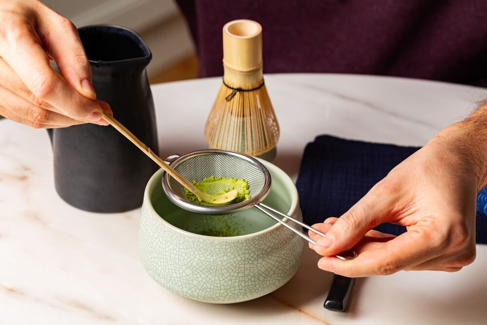 A person pressed matcha powder through a sifter into a bowl