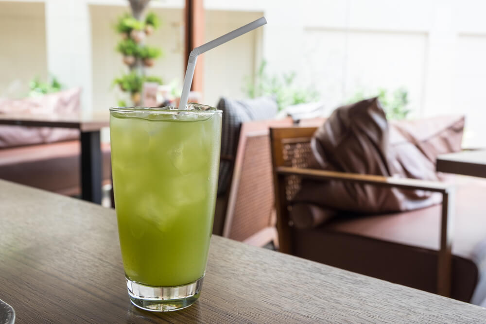 a matcha lemonade in a glass cup sits on a wooden table with a straw sticking out of it
